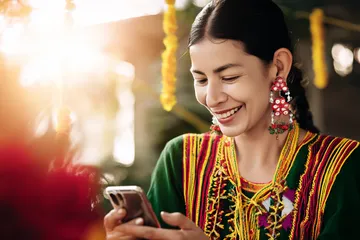 A cheerful woman in traditional clothing using her smartphone during a festive moment, showing how easily the 100JLPH app fits into everyday life.
